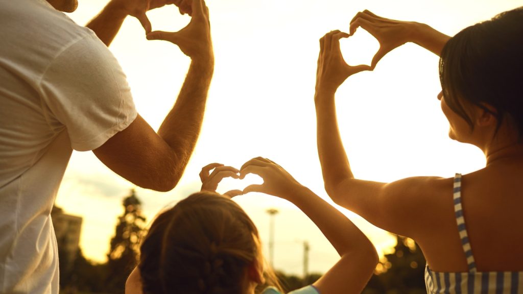 Family making heart shapes with hands at sunset. Silhouettes of three people with the sun behind them.